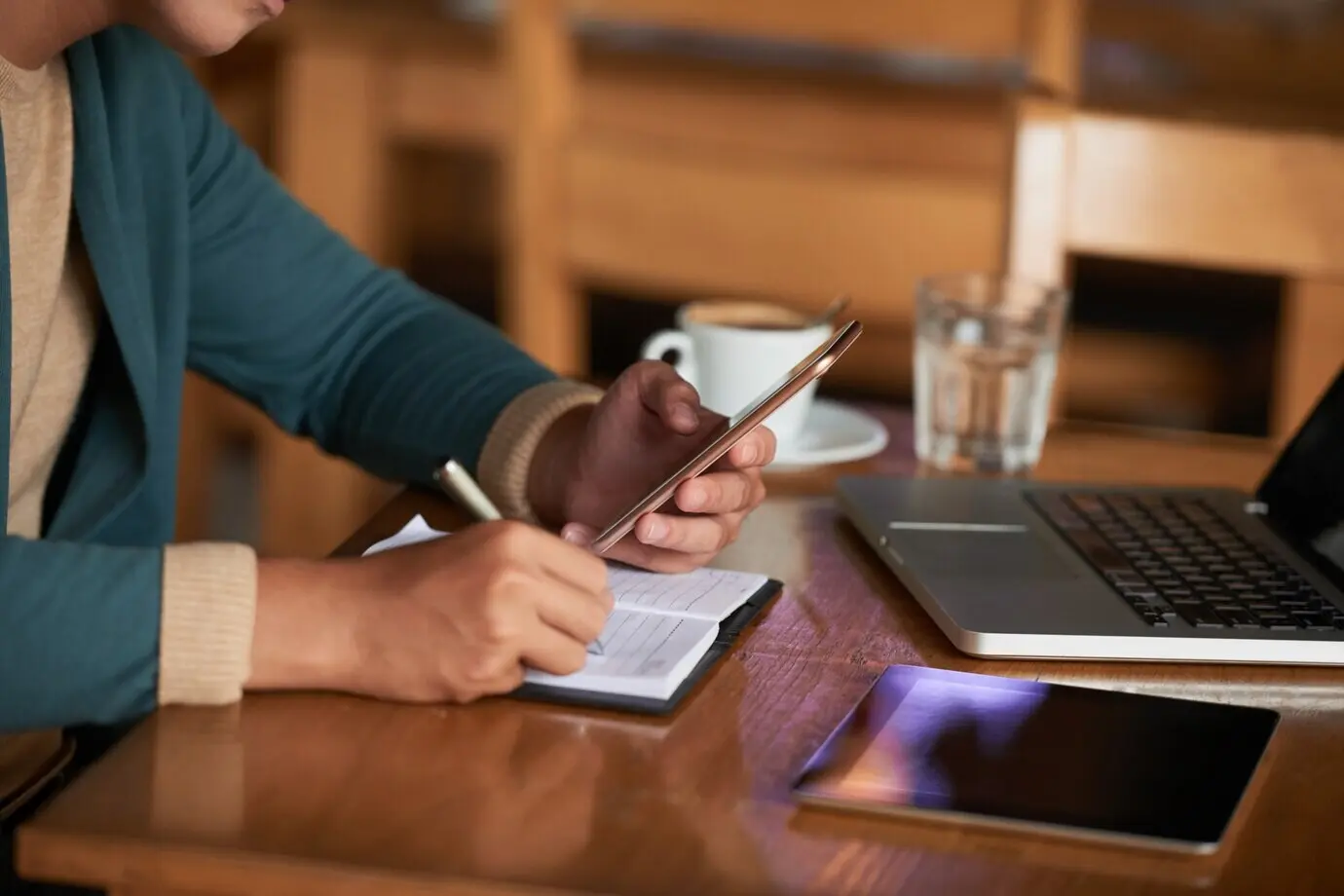 Hands of an unrecognizable man seated at a cafe table with gadgets, writing in a notebook.